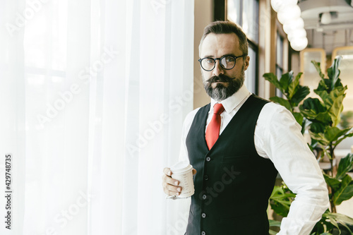 Portrait serious professional conservative adult model businessman Latin beard standing hand holding coffee cup in modern office with copy space for text.