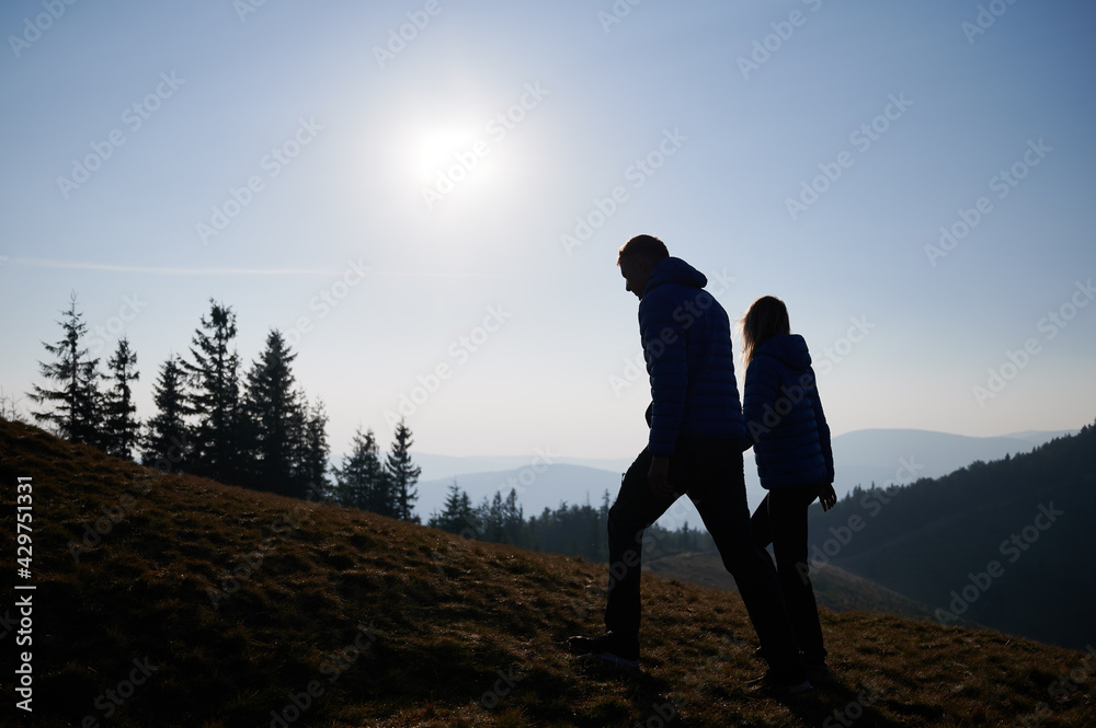 Silhouettes of love pair holding hands climbing on mountain hill, looking at surrounding landscape on bright sunny day. Active hiking day in the mountains.