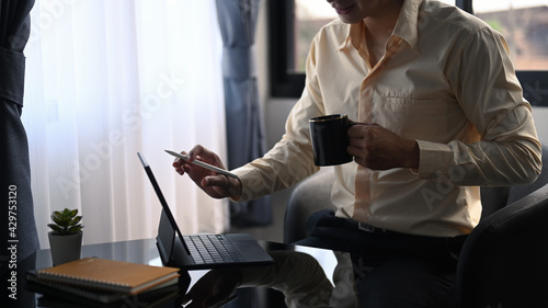 Cropped shot of happy businessman holding cup of coffee and using computer tablet in office room.