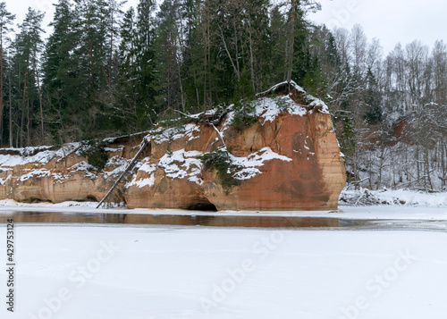 landscape with frozen river Gauja near Gudu cliffs, view of sandstone cliff from the opposite bank of the river, beautiful red rock wall, bare trees on the river bank, winter, Ligatne, Latvia photo