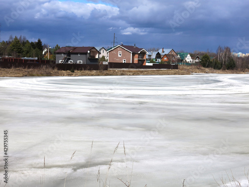 country pond, covered with a crust of spring ice with cozy houses in the distance