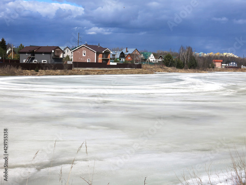 country pond, covered with a crust of spring ice with cozy houses in the distance