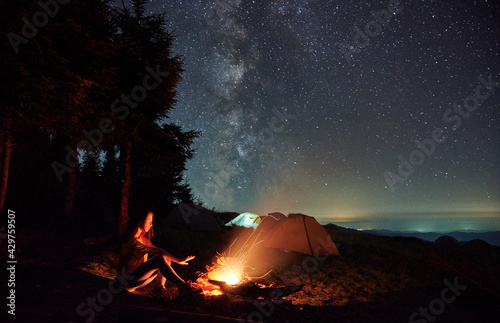 Beautiful view of night starry sky over grassy hill with illuminated camp tents and girl traveler near campfire. Young woman hiker warming hand near bonfire under magical sky with stars.