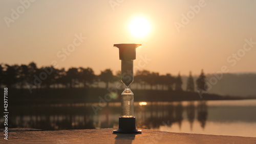 Hourglass outdoors during dawn. Start of a new cycle of life, new day. Value of time in life, an eternity. Hourglass on the background of the lake and the rising sun.
