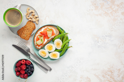 Healthy breakfast - open sandwich with salmon and cucumber, boiled eggs, peas, coffee, biscuits, nuts and berries on a bright background. Top view. Copy space
