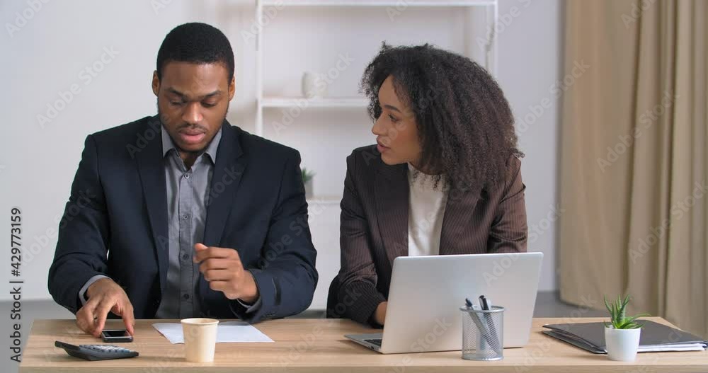 Afro american business man and woman colleague managers sitting at table working curly girl speaks secret into male ear secret tells about bad news guy feels shock urgently calls using mobile phone Afro american business man and woman colleague managers sitting at table working curly girl speaks secret into male ear secret tells about bad news guy feels shock urgently calls using mobile phone
