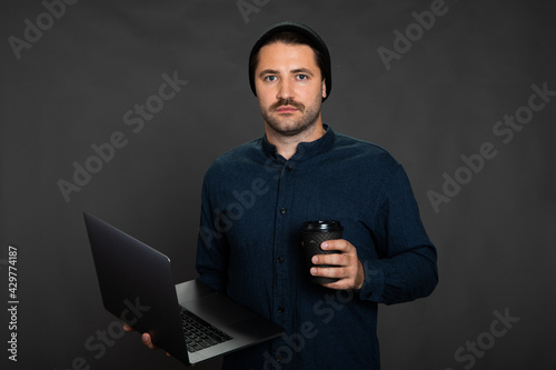 Portrait of young hipster moustached man wearing hat posing on isotaled grey background holding laptop and coffee cup. Concept of freelance work, IT professions, remote study, online education courses photo
