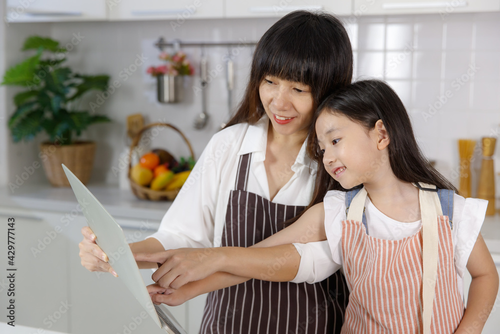 A cute and beautiful mother showing and pointing notebook computer screen for her young lovely 7 years old daughter during teaching how-to make food and cooking A cute and beautiful mother showing and pointing notebook computer screen for her young lovely 7 years old daughter during teaching how-to make food and cooking