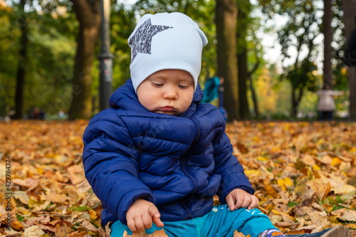  beautiful one-year-old baby is sad sitting on the grass in the autumn park with a blue cap with a star