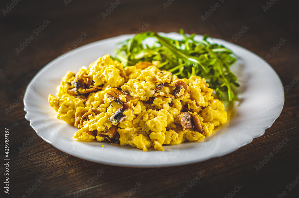 scrambled eggs with mushrooms and arugula salad in white plate on wooden table background scrambled eggs with mushrooms and arugula salad in white plate on wooden table background