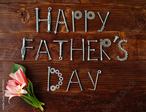 Text Happy Father's day made from metallic bolts and iron nails with flowers on wooden background. Greeting card. Creative congrats concept for dad's day celebration.