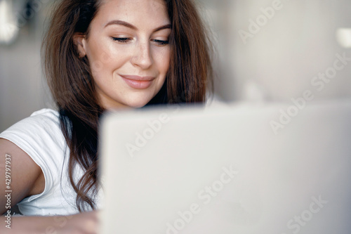 Portrait of a woman sitting on a sofa and typing text on a computer. A brunette of European appearance. Orders clothes online at home. A cozy apartment for a single strong confident woman.