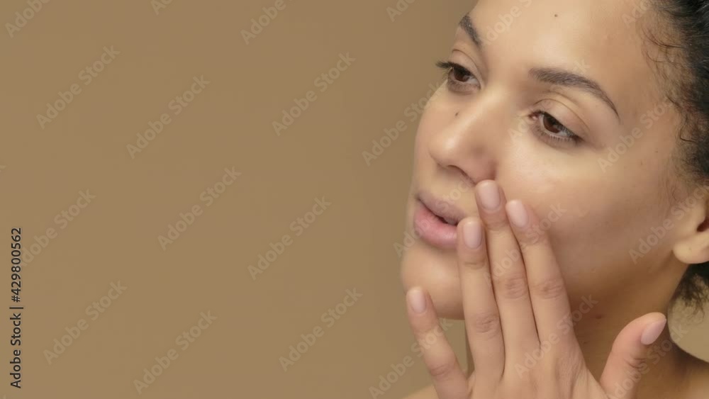 Beauty portrait young African American woman applying skincare serum on face, rubbing gently with fingers. Black female posing on brown studio background. Close up. Slow motion ready, 4K at 59.94fps. Beauty portrait young African American woman applying skincare serum on face, rubbing gently with fingers. Black female posing on brown studio background. Close up. Slow motion ready, 4K at 59.94fps.