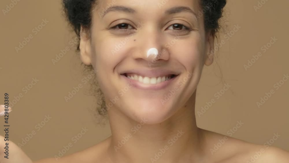 Beauty portrait of young African American woman smears face care cream on tip of her nose and smiles happily. Black female model on brown studio background. Close up. Slow motion ready, 4K at 59.94fps Beauty portrait of young African American woman smears face care cream on tip of her nose and smiles happily. Black female model on brown studio background. Close up. Slow motion ready, 4K at 59.94fps