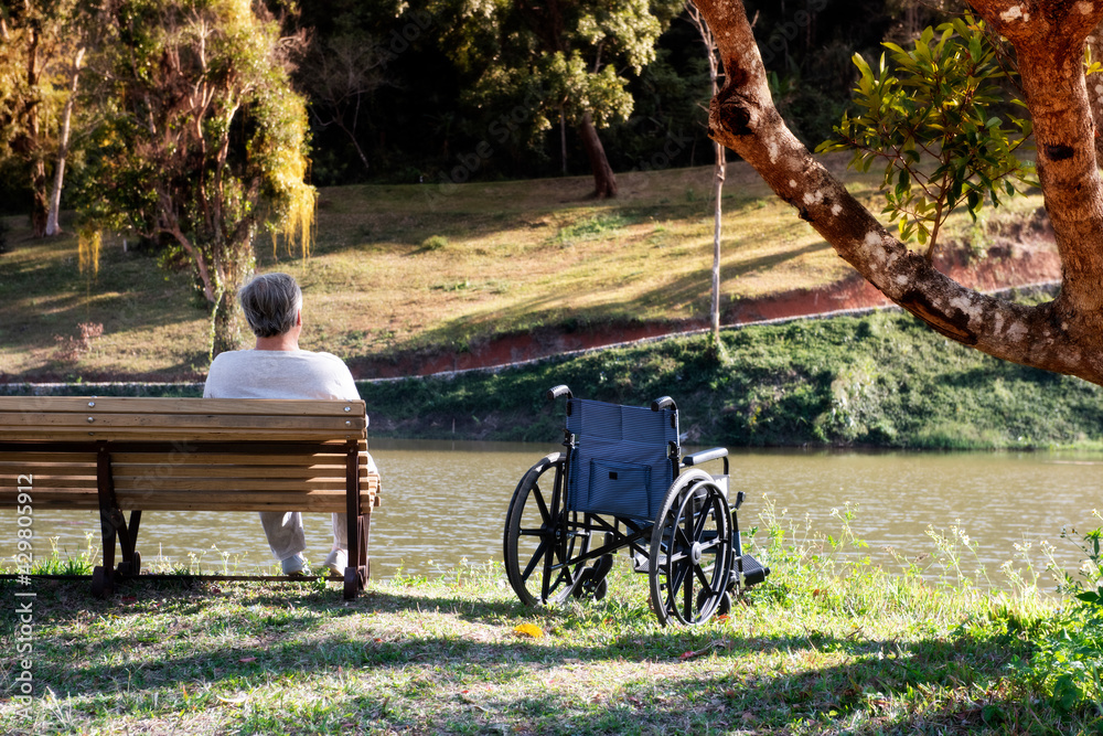 Senior is sitting alone on a wheelchair looking By the lake at evening- Special needs care assistant working with disability