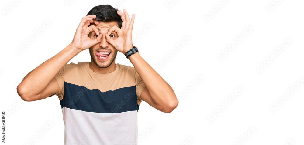 Young handsome man wearing casual clothes doing ok gesture like binoculars sticking tongue out, eyes looking through fingers. crazy expression. Young handsome man wearing casual clothes doing ok gesture like binoculars sticking tongue out, eyes looking through fingers. crazy expression.
