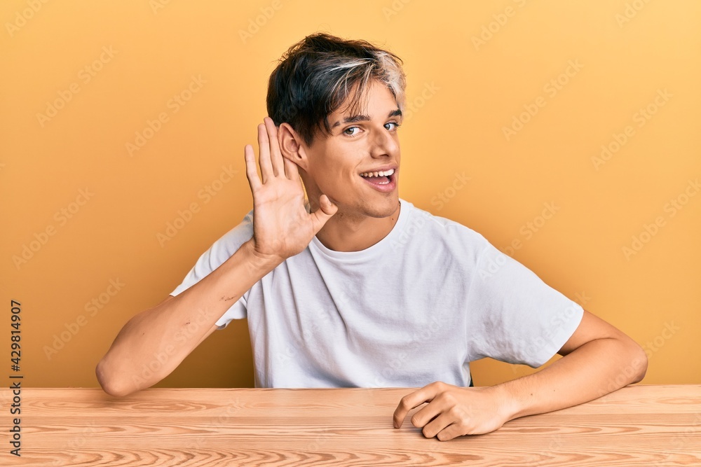 Young hispanic man wearing casual clothes sitting on the table smiling with hand over ear listening and hearing to rumor or gossip. deafness concept. Young hispanic man wearing casual clothes sitting on the table smiling with hand over ear listening and hearing to rumor or gossip. deafness concept.