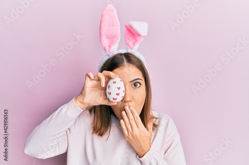 Young beautiful woman wearing cute easter bunny ears holding egg covering mouth with hand, shocked and afraid for mistake. surprised expression