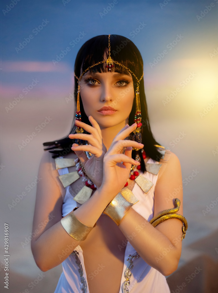 Portrait of Egypt Style woman. Sexy girl goddess Queen Cleopatra stand in  desert pyramids. Art ancient pharaoh costume white dress gold accessories.  Egyptian makeup. Creative headband, snake bracelet. Stock Photo | Adobe