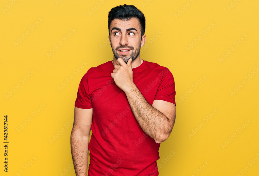 Hispanic man with beard wearing casual red t shirt with hand on chin thinking about question, pensive expression. smiling with thoughtful face. doubt concept. Hispanic man with beard wearing casual red t shirt with hand on chin thinking about question, pensive expression. smiling with thoughtful face. doubt concept.