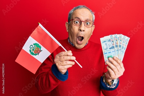 Handsome senior man with grey hair holding peru flag and peruvian sol banknotes afraid and shocked with surprise and amazed expression, fear and excited face.