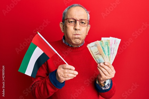 Handsome senior man with grey hair holding united arab emirates dirham banknotes puffing cheeks with funny face. mouth inflated with air, catching air.