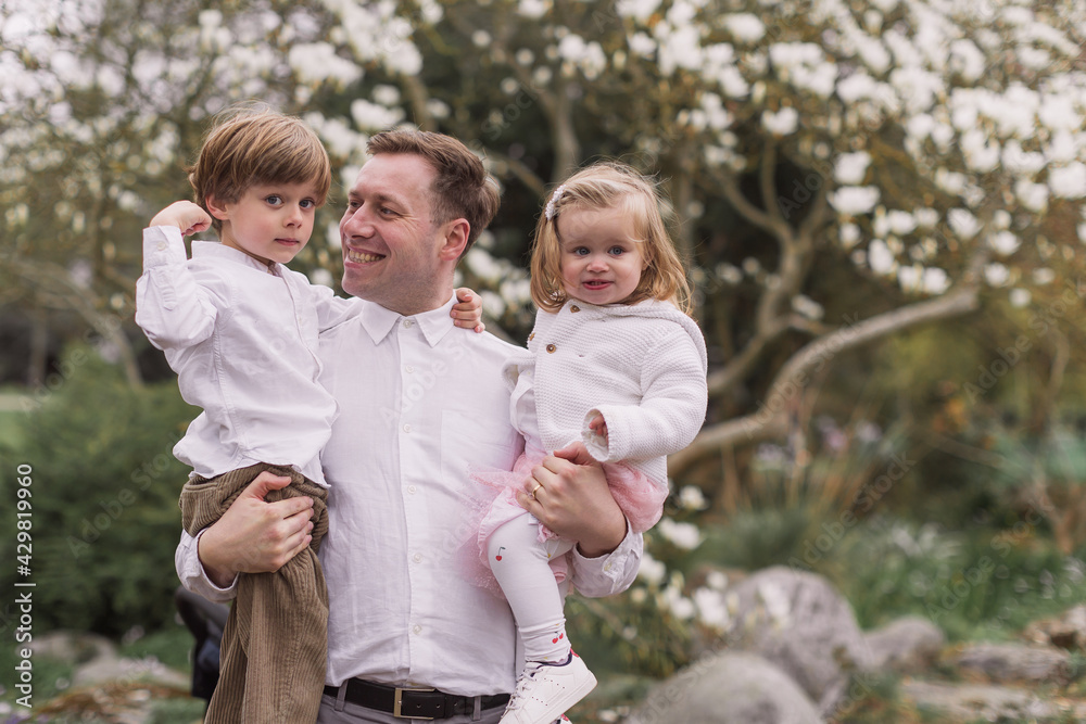 Fototapeta premium Happy young father holding daughter and son in his arms in the spring park. Standing. Smiling. Background of blooming magnolia.