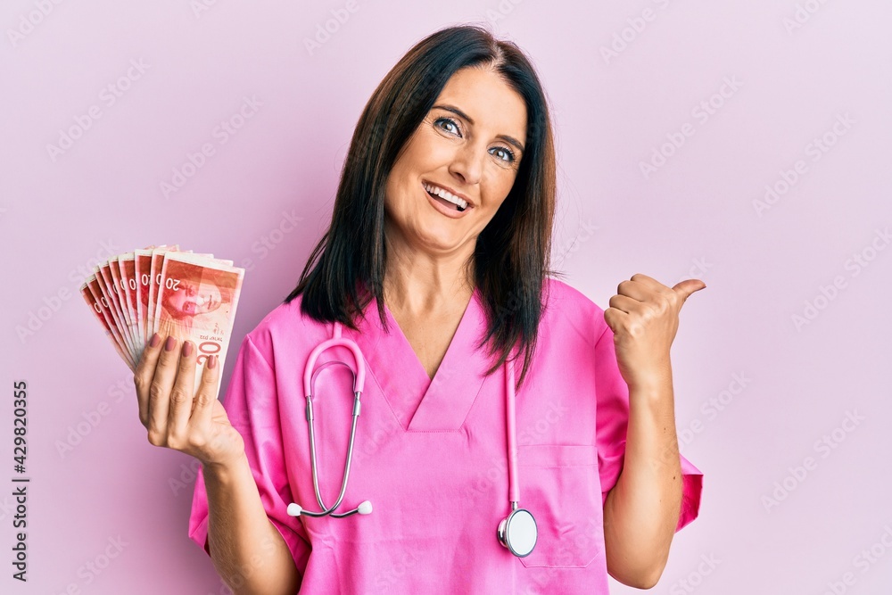 Middle age brunette doctor woman holding 20 shekels banknotes pointing thumb up to the side smiling happy with open mouth Middle age brunette doctor woman holding 20 shekels banknotes pointing thumb up to the side smiling happy with open mouth