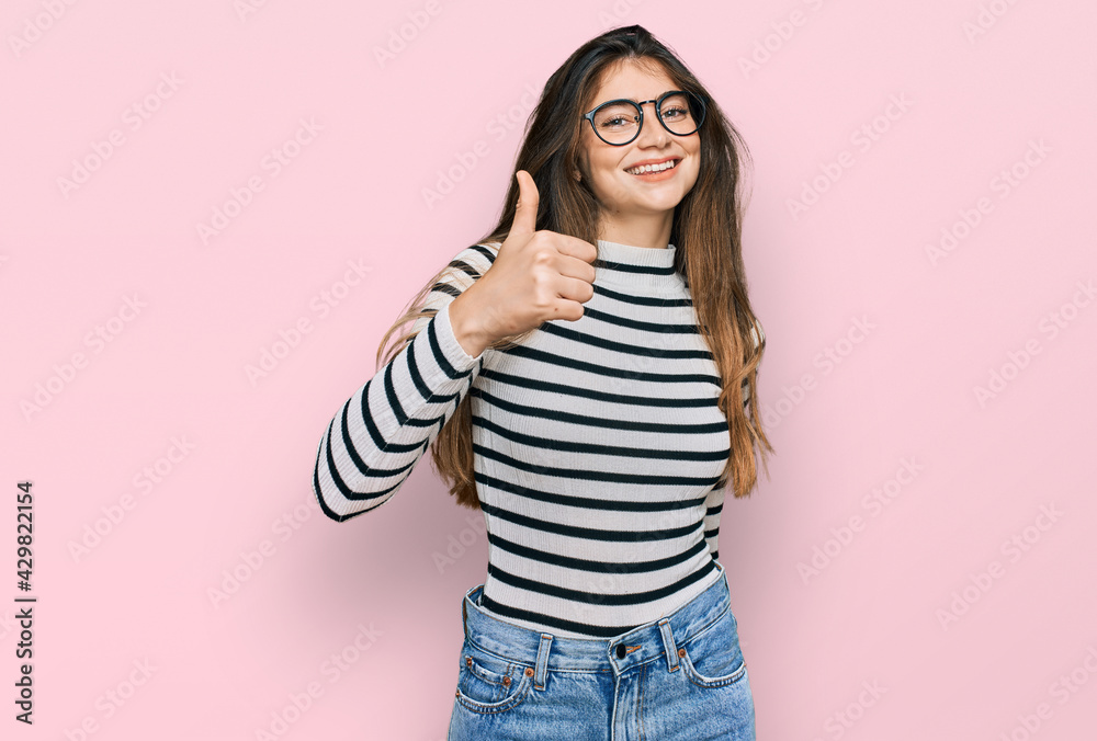 Young beautiful teen girl wearing casual clothes and glasses doing happy thumbs up gesture with hand. approving expression looking at the camera showing success. Young beautiful teen girl wearing casual clothes and glasses doing happy thumbs up gesture with hand. approving expression looking at the camera showing success.