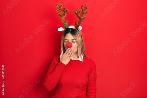 Beautiful hispanic woman wearing deer christmas hat and red nose bored yawning tired covering mouth with hand. restless and sleepiness.