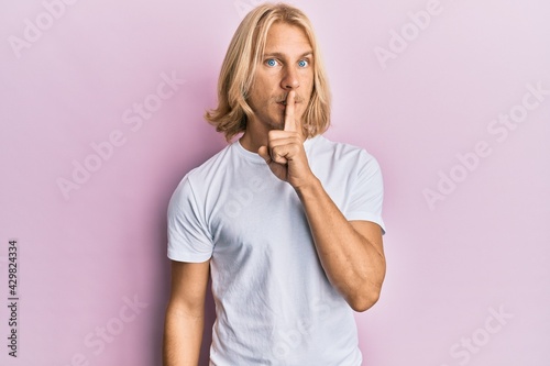 Caucasian young man with long hair wearing casual white t shirt asking to be quiet with finger on lips. silence and secret concept.