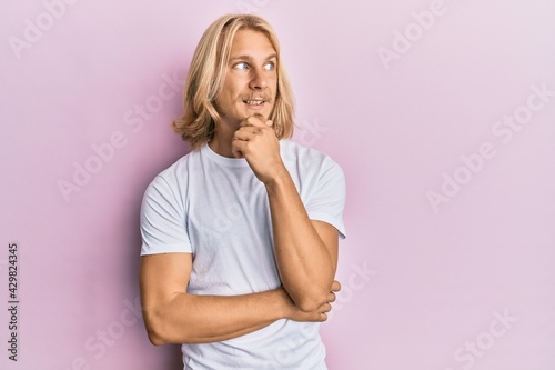 Caucasian young man with long hair wearing casual white t shirt with hand on chin thinking about question, pensive expression. smiling with thoughtful face. doubt concept.