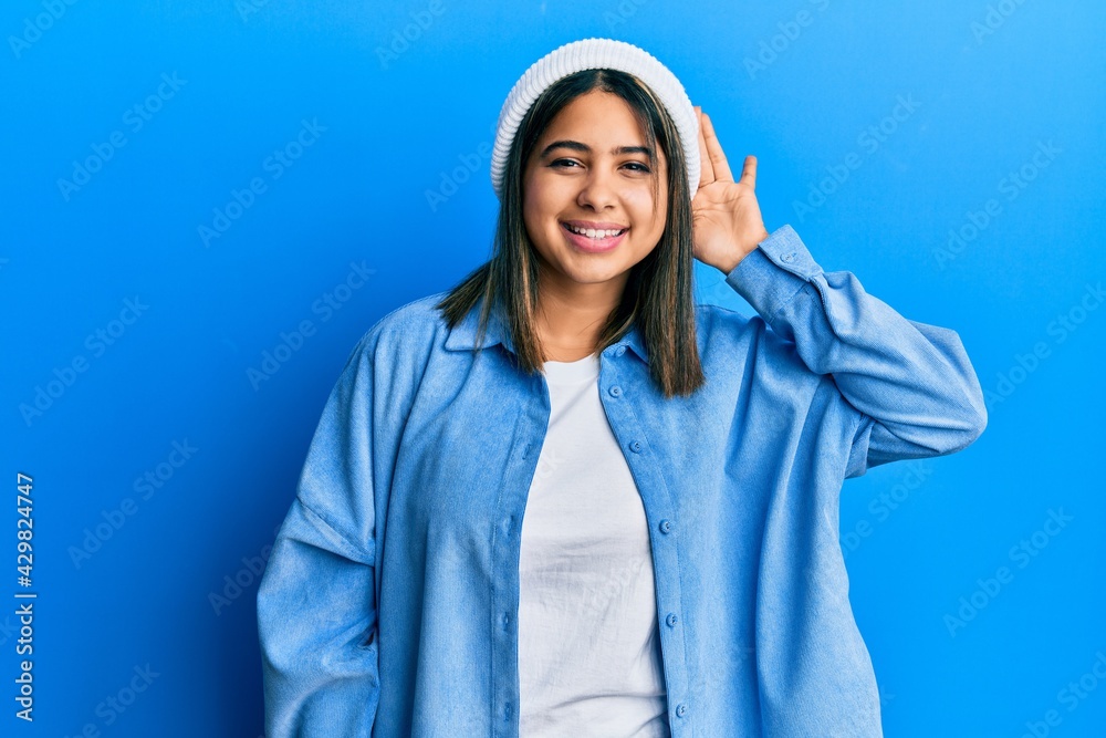 Young latin woman wearing cute wool cap smiling with hand over ear listening an hearing to rumor or gossip. deafness concept. Young latin woman wearing cute wool cap smiling with hand over ear listening an hearing to rumor or gossip. deafness concept.