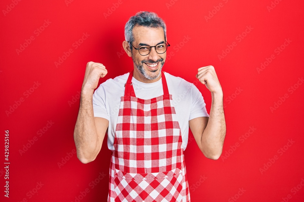 Naklejka premium Handsome middle age man with grey hair wearing apron screaming proud, celebrating victory and success very excited with raised arms
