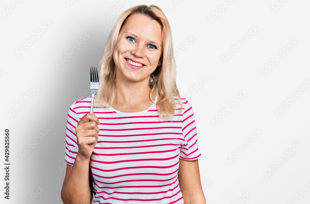 Young caucasian woman holding one silver fork looking positive and happy standing and smiling with a confident smile showing teeth Young caucasian woman holding one silver fork looking positive and happy standing and smiling with a confident smile showing teeth