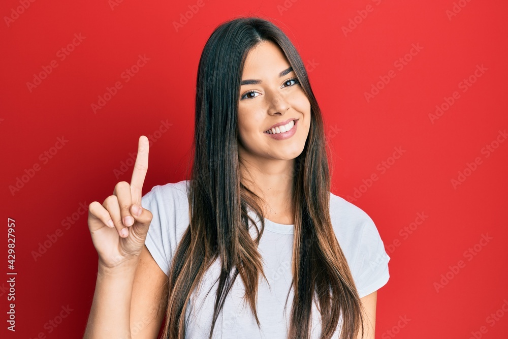 Young brunette woman wearing casual white tshirt over red background showing and pointing up with finger number one while smiling confident and happy. Young brunette woman wearing casual white tshirt over red background showing and pointing up with finger number one while smiling confident and happy.