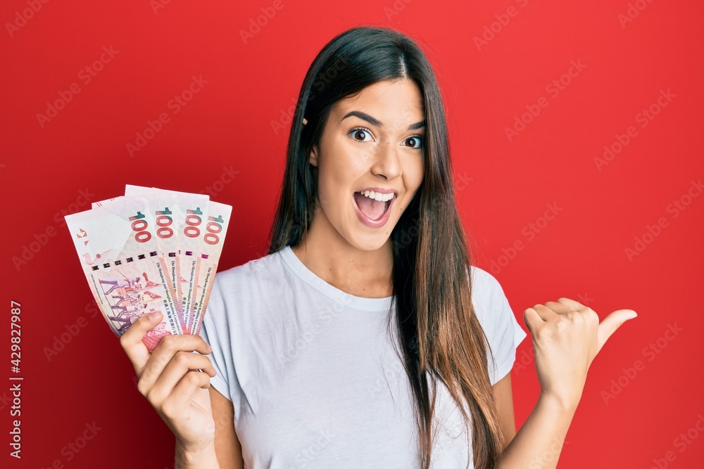 Young brunette woman holding hong kong 100 dollars banknotes pointing thumb up to the side smiling happy with open mouth Young brunette woman holding hong kong 100 dollars banknotes pointing thumb up to the side smiling happy with open mouth