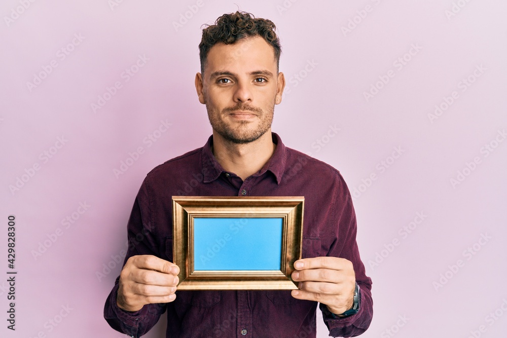 Young hispanic man holding empty frame relaxed with serious expression on face. simple and natural looking at the camera.
