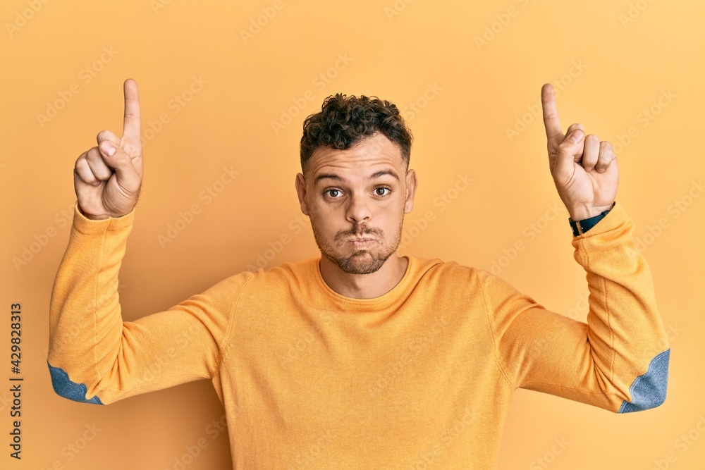 Young hispanic man pointing up with fingers puffing cheeks with funny face. mouth inflated with air, catching air. Young hispanic man pointing up with fingers puffing cheeks with funny face. mouth inflated with air, catching air.