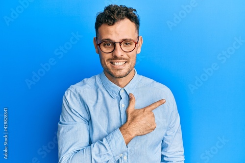 Young hispanic man wearing casual clothes and glasses cheerful with a smile on face pointing with hand and finger up to the side with happy and natural expression