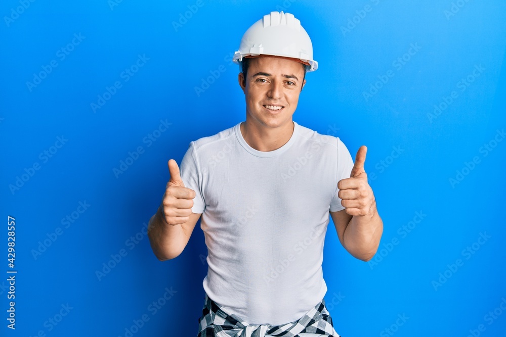 Handsome young man wearing builder uniform and hardhat success sign doing positive gesture with hand, thumbs up smiling and happy. cheerful expression and winner gesture.