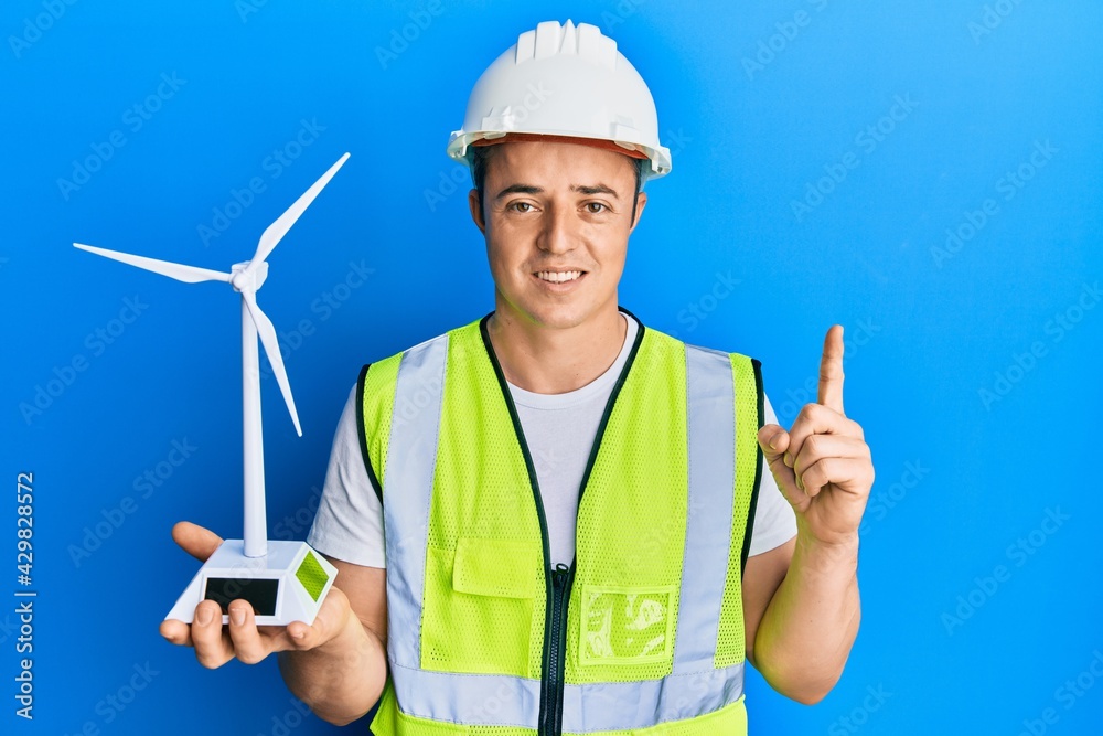 Handsome young man holding solar windmill for renewable electricity smiling with an idea or question pointing finger with happy face, number one