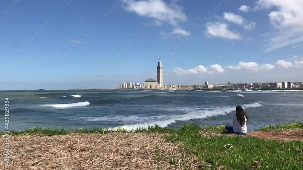 Casablanca, Morocco - March 14 2021 : Beautiful free and wild woman, sitting in meditation pose on the Casablanca beach in front of the Hassan II mosque