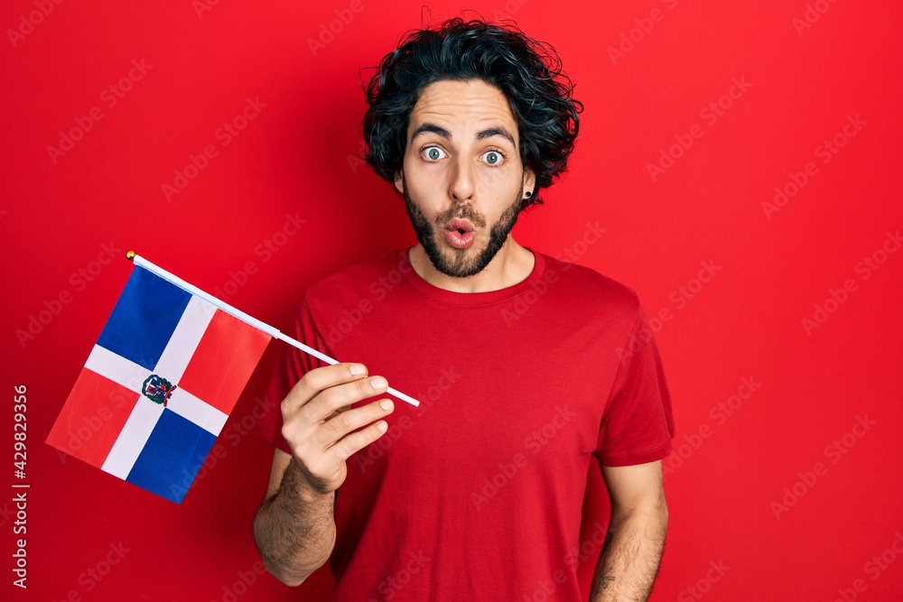 Handsome hispanic man holding dominican republic flag scared and amazed with open mouth for surprise, disbelief face Handsome hispanic man holding dominican republic flag scared and amazed with open mouth for surprise, disbelief face