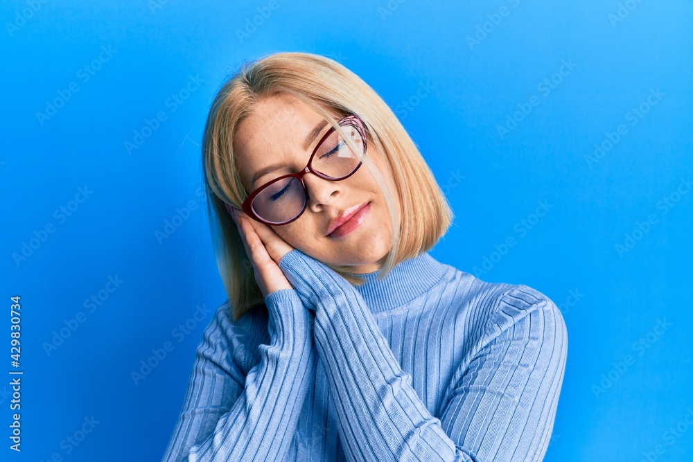 Young blonde woman wearing casual clothes and glasses sleeping tired dreaming and posing with hands together while smiling with closed eyes. Young blonde woman wearing casual clothes and glasses sleeping tired dreaming and posing with hands together while smiling with closed eyes.