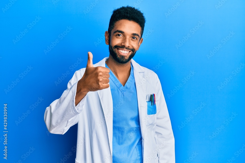Handsome hispanic man with beard wearing doctor uniform doing happy thumbs up gesture with hand. approving expression looking at the camera showing success. Handsome hispanic man with beard wearing doctor uniform doing happy thumbs up gesture with hand. approving expression looking at the camera showing success.
