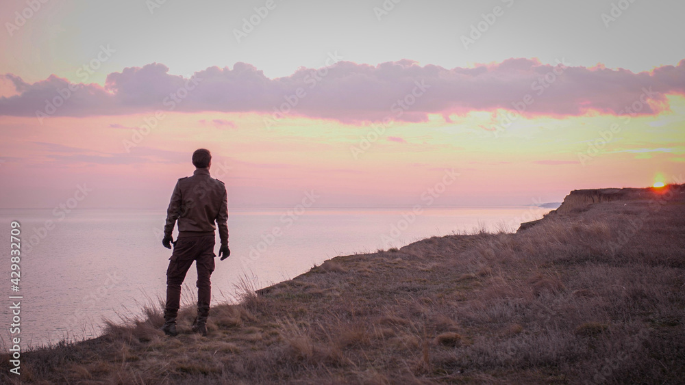 Looking on young biker in leather suit stay on a precipice above sea looking far on majestic sunset with rose clouds. Looking on young biker in leather suit stay on a precipice above sea looking far on majestic sunset with rose clouds.