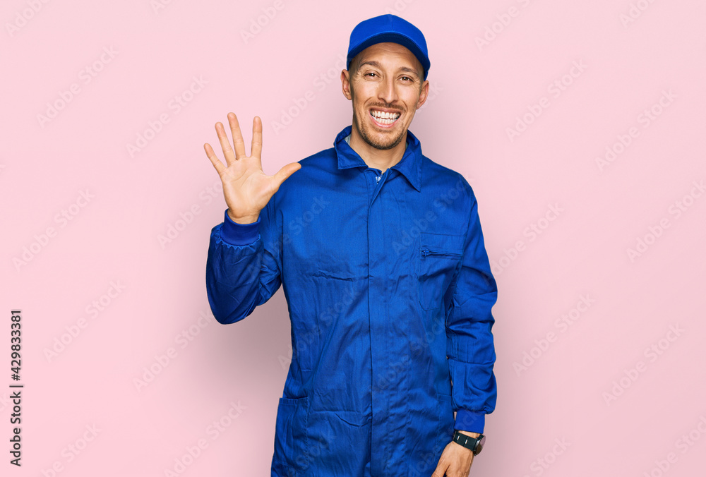 Bald man with beard wearing builder jumpsuit uniform showing and pointing up with fingers number five while smiling confident and happy. Bald man with beard wearing builder jumpsuit uniform showing and pointing up with fingers number five while smiling confident and happy.