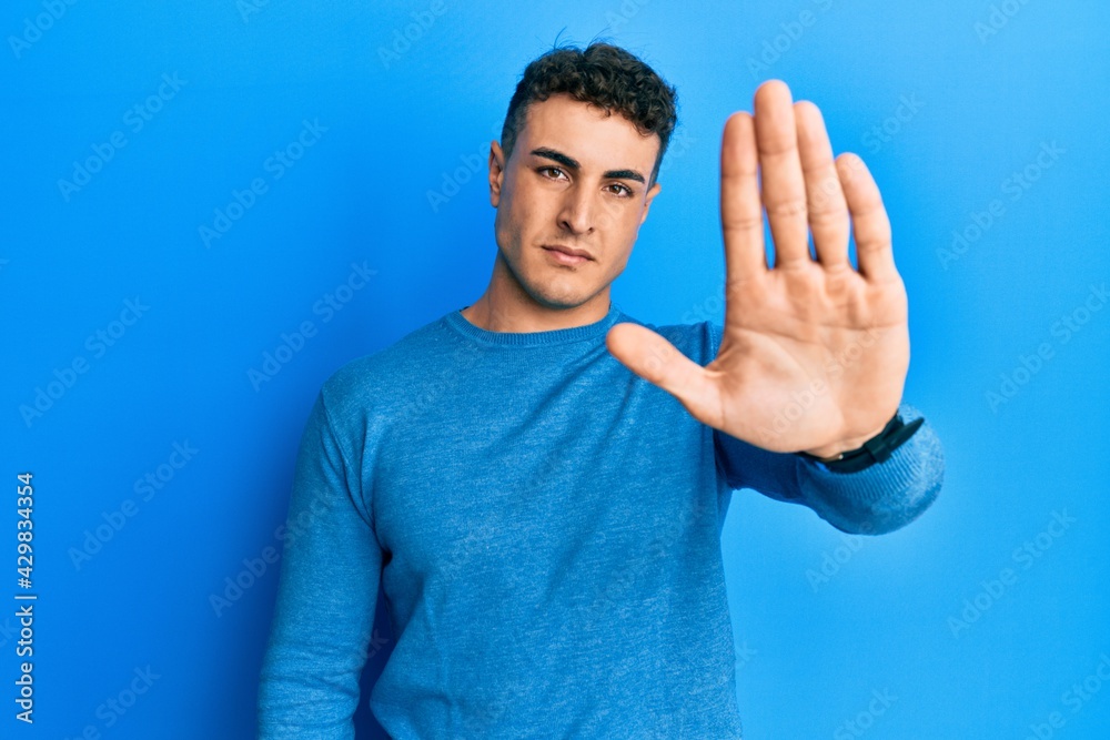 Naklejka premium Hispanic young man wearing casual winter sweater doing stop sing with palm of the hand. warning expression with negative and serious gesture on the face.