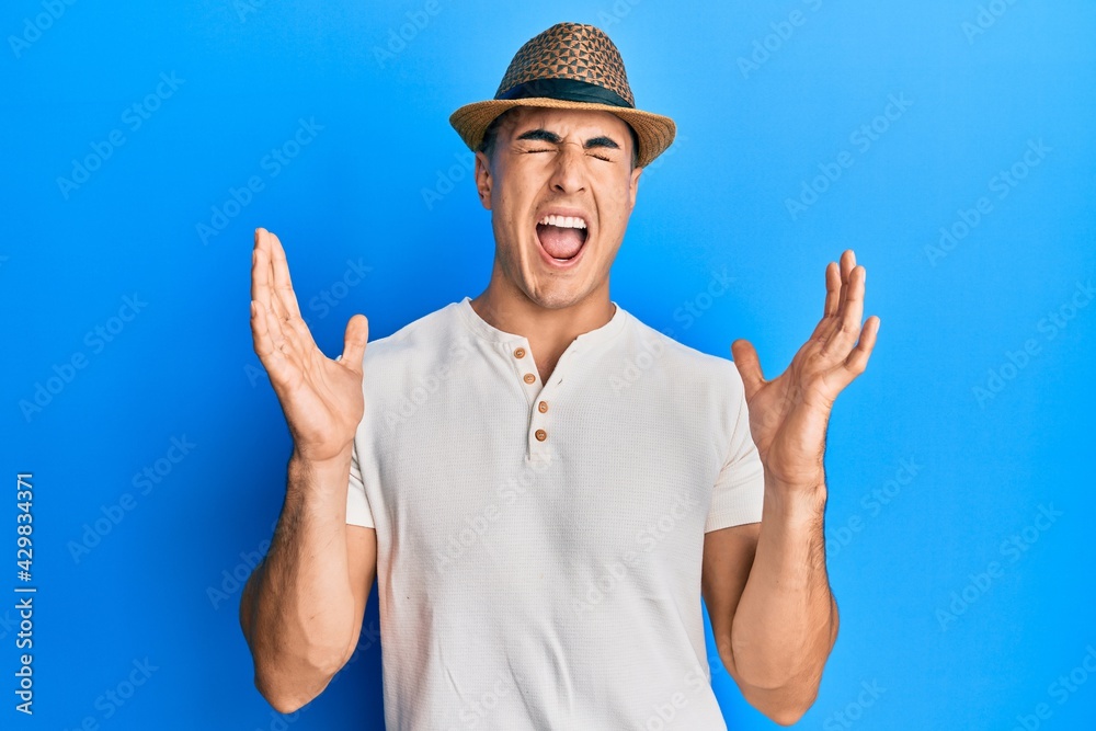 Hispanic young man wearing summer hat celebrating mad and crazy for success with arms raised and closed eyes screaming excited. winner concept Hispanic young man wearing summer hat celebrating mad and crazy for success with arms raised and closed eyes screaming excited. winner concept